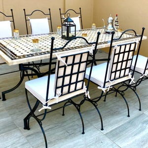 Op de afbeelding: Een buiteneetset met een rechthoekige tafel met een moza&iuml;ektegelblad en zwarte metalen stoelen. Zes bijpassende stoelen met witte kussens en rasterruggen omringen de tafel. Decoratieve items, waaronder glazen en een lantaarn, staan op de tafel.
