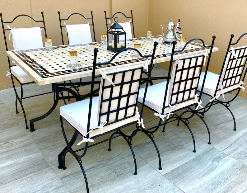 Op de afbeelding: Een buiteneetset met een rechthoekige tafel met een moza&iuml;ektegelblad en zwarte metalen stoelen. Zes bijpassende stoelen met witte kussens en rasterruggen omringen de tafel. Decoratieve items, waaronder glazen en een lantaarn, staan op de tafel.
