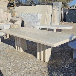 Peut inclure: Une grande table rectangulaire en pierre beige avec une surface polie. La table est soutenue par quatre pieds cannelés épais. Plusieurs autres tables et dalles en pierre sont visibles en arrière-plan, suggérant un fournisseur ou une salle d'exposition de pierre.