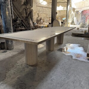 May include: A large rectangular marble table with a decorative border. The table is supported by three cylindrical, fluted legs. The table appears to be in a workshop or studio setting.