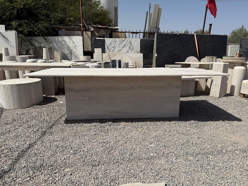 Pu&ograve; includere: Un grande tavolo rettangolare in pietra di colore beige chiaro. Il tavolo ha una base solida e un piano piatto. Sullo sfondo sono visibili altre lastre di pietra e strutture cilindriche, che suggeriscono un fornitore o un laboratorio di pietra.