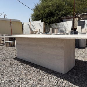 Pu&ograve; includere: Un grande tavolo rettangolare in pietra di colore beige chiaro. Il tavolo ha una base solida a forma di blocco e un piano piatto. Si trova all'aperto, tra ghiaia e varie lastre di pietra. Il tavolo &egrave; lungo circa 200 cm.