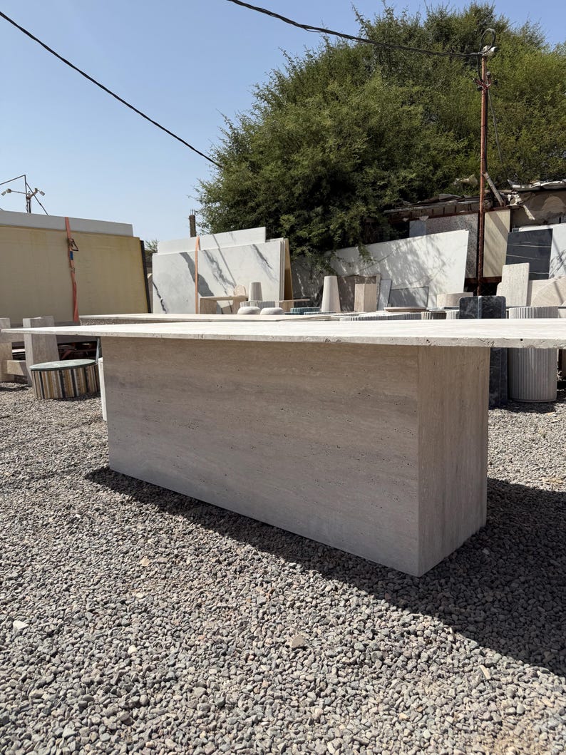 Pu&ograve; includere: Un grande tavolo rettangolare in pietra di colore beige chiaro. Il tavolo ha una base solida a forma di blocco e un piano piatto. Si trova all'aperto, tra ghiaia e varie lastre di pietra. Il tavolo &egrave; lungo circa 200 cm.