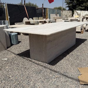 Pu&ograve; includere: Un grande tavolo rettangolare in pietra di colore beige chiaro. Il tavolo ha una base spessa e solida e un piano liscio e piatto. &Egrave; circondato da altre lastre e tavoli in pietra, suggerendo un cortile di pietra o un'esposizione di mobili da esterno. Misura circa 300 cm di lunghezza.
