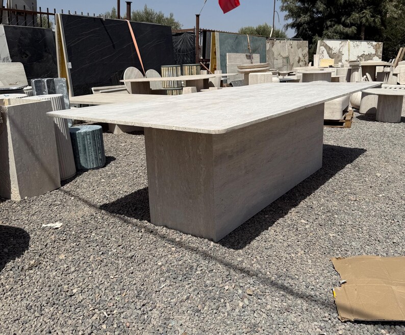 Pu&ograve; includere: Un grande tavolo rettangolare in pietra di colore beige chiaro. Il tavolo ha una base spessa e solida e un piano liscio e piatto. &Egrave; circondato da altre lastre e tavoli in pietra, suggerendo un cortile di pietra o un'esposizione di mobili da esterno. Misura circa 300 cm di lunghezza.