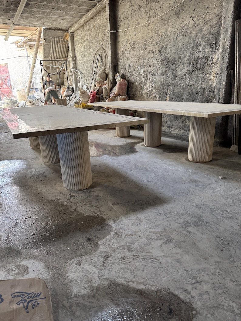 May include: Two rectangular tables with light-colored tops and cylindrical, fluted legs. The tables are in a workshop setting with a concrete floor and a person in the background. The tables appear to be made of stone or a similar material.