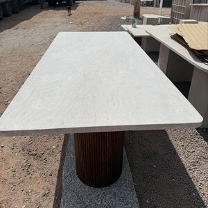 May include: A rectangular stone table with a light grey top and a dark brown, cylindrical base. The base has a vertical, ribbed design. The table is outdoors on a gravel surface.