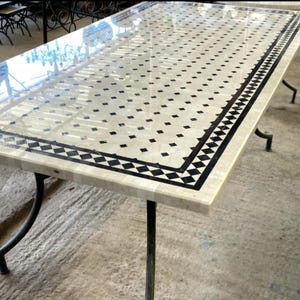 Op de afbeelding: Rechthoekige tafel met een cr&egrave;mekleurig oppervlak, versierd met een patroon van zwarte vierkanten en een zwart-witte rand. De tafel wordt ondersteund door zwarte metalen poten, die een mix van klassieke en moderne designelementen laten zien. De lengte is ongeveer 180 cm.