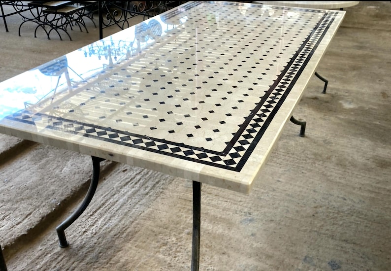 Op de afbeelding: Rechthoekige tafel met een cr&egrave;mekleurig oppervlak, versierd met een patroon van zwarte vierkanten en een zwart-witte rand. De tafel wordt ondersteund door zwarte metalen poten, die een mix van klassieke en moderne designelementen laten zien. De lengte is ongeveer 180 cm.