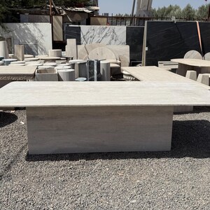 Pu&ograve; includere: Un grande tavolo rettangolare in pietra di colore beige chiaro. Il tavolo ha una base solida e un piano piatto. Altri tavoli in pietra ed elementi architettonici sono visibili sullo sfondo.
