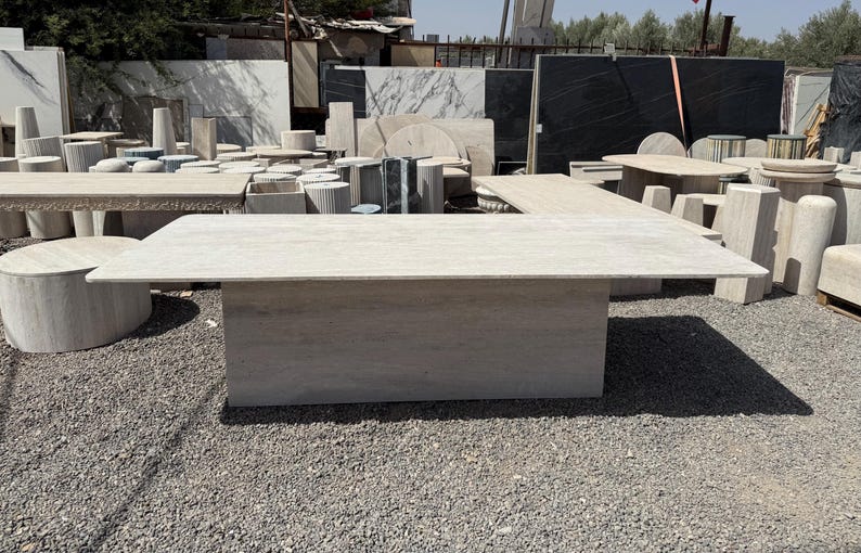 Pu&ograve; includere: Un grande tavolo rettangolare in pietra di colore beige chiaro. Il tavolo ha una base solida e un piano piatto. Altri tavoli in pietra ed elementi architettonici sono visibili sullo sfondo.