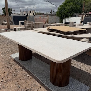 Peut inclure: Une table rectangulaire en pierre avec un plateau clair et deux supports cylindriques en bois brun foncé. La table repose sur une base en pierre grise, présentant un design moderne pour l'intérieur ou l'extérieur.