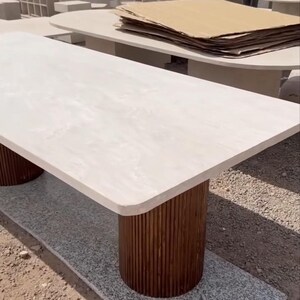 May include: A rectangular table with a light-coloured stone top and two cylindrical, dark brown wooden legs. The table is set on a grey surface, with other tables visible in the background. The table's dimensions are not visible.