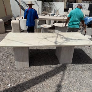 Peut inclure: Une grande table rectangulaire en pierre de couleur beige clair. La table a une surface lisse et est soutenue par deux socles rectangulaires. La table mesure environ 244 cm de long et 122 cm de large. D'autres tables et dalles en pierre sont visibles en arrière-plan.