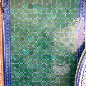 May include: A wall covered in green and blue tiles. The tiles are square with a diamond-shaped blue accent in the center of each. A blue border frames the tiled wall. The tiles are a mix of light and dark green.