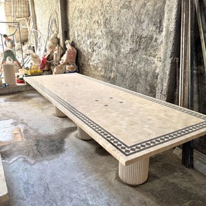 May include: A large rectangular mosaic table with a beige and black geometric border. The table is supported by four cylindrical legs. The table measures approximately 120 inches long.