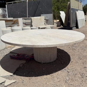 Könnte beinhalten: Ein großer, runder Steintisch mit hellbeiger Oberfläche. Der Tisch hat ein zylindrisches Untergestell aus vertikalen Steinelementen. Der Tisch befindet sich im Freien, mit anderen Steinplatten und Materialien im Hintergrund.