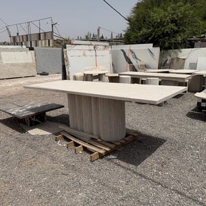 Puede incluir: Una mesa grande y rectangular de piedra con una tapa beige claro y una base cilíndrica. La base está hecha de piezas de piedra verticales y texturizadas. La mesa está sobre un palet de madera en un entorno al aire libre, con otras losas de piedra al fondo.