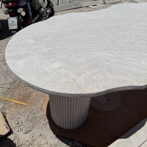 Peut inclure: Une table de couleur gris clair, de forme organique, avec un pied cylindrique. Le plateau a une surface lisse et texturée. Le pied est composé de tiges verticales gris clair. La table est probablement en pierre ou en béton.