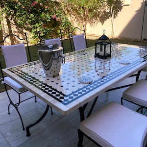 Op de afbeelding: Eettafel voor buiten met een moza&iuml;ekblad met zwart-witte geometrische patronen. De tafel heeft een zwart metalen frame en is omringd door stoelen met witte kussens. Een zilveren ijsemmer en een zwarte lantaarn staan op de tafel.