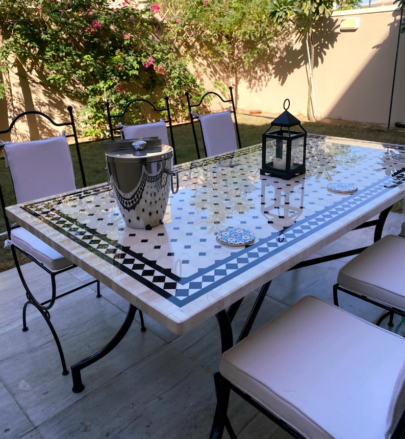 Op de afbeelding: Eettafel voor buiten met een moza&iuml;ekblad met zwart-witte geometrische patronen. De tafel heeft een zwart metalen frame en is omringd door stoelen met witte kussens. Een zilveren ijsemmer en een zwarte lantaarn staan op de tafel.