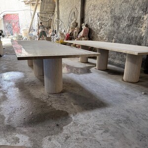 May include: Two large, light-colored rectangular tables with thick, cylindrical legs. The tables are in a workshop setting with a concrete floor and walls. The tables appear to be made of wood or a similar material.