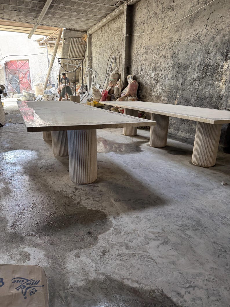 May include: Two large, light-colored rectangular tables with thick, cylindrical legs. The tables are in a workshop setting with a concrete floor and walls. The tables appear to be made of wood or a similar material.
