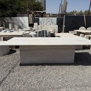 Pu&ograve; includere: Un grande tavolo rettangolare in pietra di colore grigio chiaro. Il tavolo ha una base solida e un piano piatto. Altri tavoli e lastre di pietra sono visibili sullo sfondo, suggerendo un fornitore o un laboratorio di pietra.