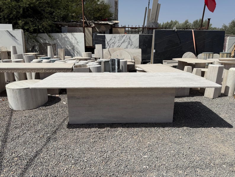 Pu&ograve; includere: Un grande tavolo rettangolare in pietra di colore grigio chiaro. Il tavolo ha una base solida e un piano piatto. Altri tavoli e lastre di pietra sono visibili sullo sfondo, suggerendo un fornitore o un laboratorio di pietra.