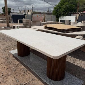 May include: A rectangular stone table with a light-coloured top and two cylindrical, dark brown wooden supports. The table rests on a grey stone base, suitable for indoor or outdoor use. The table's design is modern.