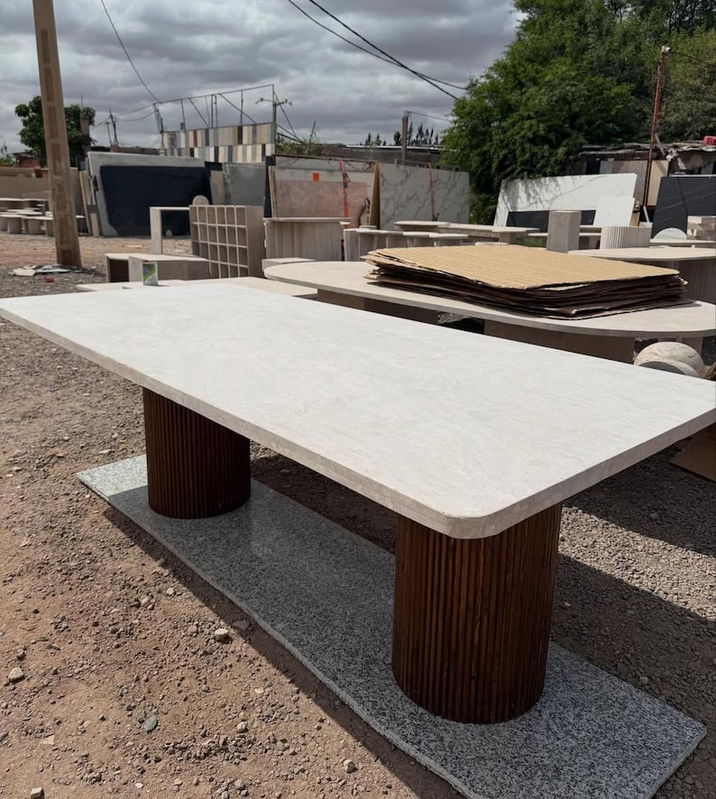 May include: A rectangular stone table with a light-coloured top and two cylindrical, dark brown wooden supports. The table rests on a grey stone base, suitable for indoor or outdoor use. The table's design is modern.