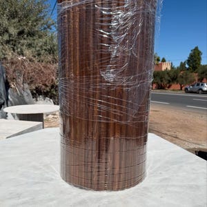 May include: A cylindrical wooden product, wrapped in clear plastic, stands on a white surface. The wood has a rich, dark brown colour with vertical grain patterns. The product appears to be new, possibly for construction or interior design.
