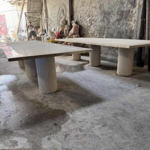 May include: Two rectangular tables with light-colored tops and cylindrical, fluted legs. The tables are in a workshop setting with a concrete floor and a person in the background. The tables appear to be made of stone or a similar material.