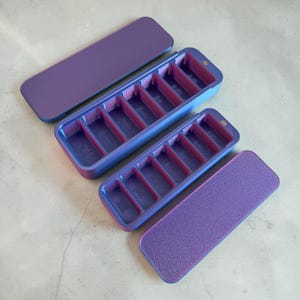 Può includere: Due portapillole rettangolari aperti con coperchi. Gli organizzatori sono divisi in sei scomparti. L'esterno è sfumato blu e viola, con interno rosa. I coperchi sono viola unito. Su una superficie grigio chiaro.