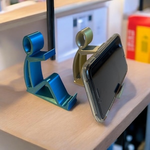 May include: Three phone stands in the shape of stylized figures. One is blue, one is gold, and the third holds a black smartphone. The stands are on a light-colored wooden surface.