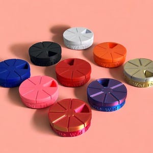 May include: A selection of multi-coloured, circular pill organisers. Each organiser is divided into sections and labelled with days of the week. The colours include blue, pink, red, and gold. The organisers are displayed on a pink surface.
