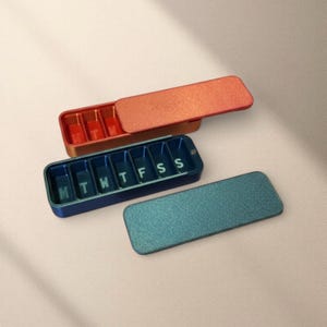 Può includere: Tre portapillole rettangolari nei toni dell'arancione, blu e verde acqua. L'organizer blu ha i giorni della settimana contrassegnati con le lettere L, M, M, G, V, S, D. L'organizer arancione è aperto, rivelando scomparti rossi.