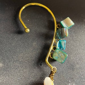 May include: A gold ear cuff with a dangling seashell charm. The ear cuff is decorated with several small, iridescent green and blue shell pieces.