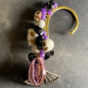 May include: A gold ear cuff with a pink seashell charm and multiple black and white skull beads, purple beads, and black beads.