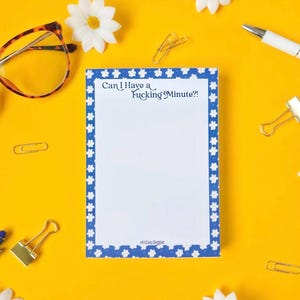 May include: A rectangular notepad with a blue border featuring a white flower pattern. The top of the page displays the text Can I Have a Fucking Minute?! in a dark blue font. The notepad is surrounded by tortoiseshell glasses and paper clips.