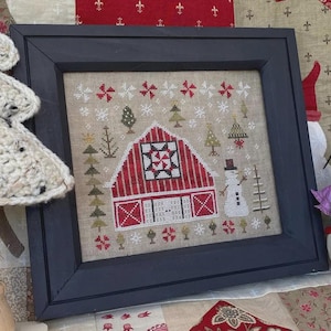 Può includere: Opera d'arte a punto croce incorniciata con un fienile rosso con un motivo a stella, pupazzi di neve e alberi di Natale. L'opera d'arte è ambientata su uno sfondo beige con fiocchi di neve ed è incorniciata in una cornice di legno blu scuro. Il tema generale è una scena di festa invernale.