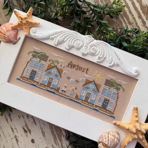 Op de afbeelding: Een ingelijst kruissteekwerk met een kustlandschap met blauwe huizen, palmbomen en het woord "August". De witte lijst is versierd met decoratieve elementen, schelpen en zeesterren, wat een strandthema oproept.