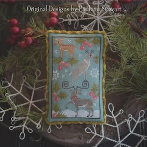 Può includere: Un ornamento natalizio rettangolare ricamato a punto croce con tre cervi, bacche e fiocchi di neve su sfondo azzurro. L'ornamento è incorniciato da un bordo giallo-verde e il testo "Original Designs by Paulette Stewart" è in alto.