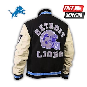 Puede incluir: Chaqueta universitaria de los Detroit Lions en negro y crema. La chaqueta presenta el logotipo del equipo y las palabras "DETROIT LIONS" en la espalda. Las mangas son color crema y tiene detalles en azul y negro.