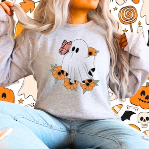 Op de afbeelding: Een lichtgrijs sweatshirt met een grillig spookontwerp. Het spook is versierd met een roze vlinder en oranje bloemen. De achtergrond is een Halloween-patroon met pompoenen, snoep en geesten.