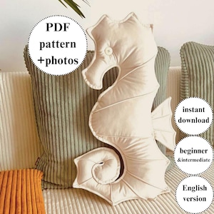 Könnte beinhalten: Ein cremefarbenes, seepferdchenförmiges Kissen mit Knopfauge und gestickten Details. Das Kissen liegt auf einem Sofa mit grünen und orangefarbenen Kissen. Text auf dem Bild: "PDF pattern + photos", "instant download", "beginner & intermediate" und "English version".