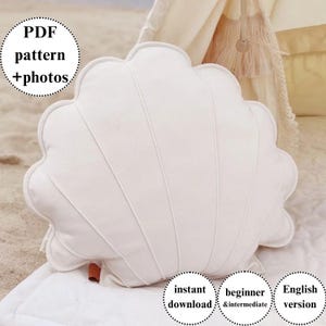 Peut inclure: Un coussin blanc en forme de coquillage avec un bord festonné et des lignes verticales cousues. Le coussin est posé sur une couverture blanche. Le texte sur l'image comprend "PDF pattern + photos", "instant download", "beginner & intermediate" et "English version".