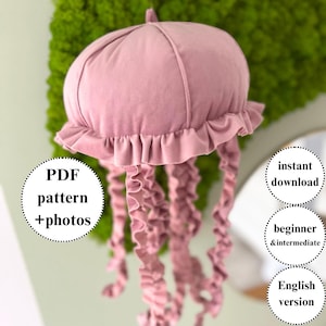 Può includere: Un oggetto decorativo a forma di medusa rosa in peluche, con un bordo a balze e lunghi tentacoli ricci. Il testo sull'immagine dice "PDF pattern + photos", "instant download", "beginner & intermediate" e "English version".