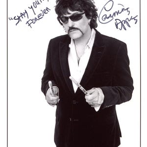 Puede incluir: Una fotografía en blanco y negro de Carmine Appice con un blazer oscuro, camisa blanca y gafas de sol, sosteniendo dos baquetas. La imagen presenta el texto manuscrito "STAY YOUNG FOREVER" y "LOVE Carmine Appice" con el nombre impreso abajo.