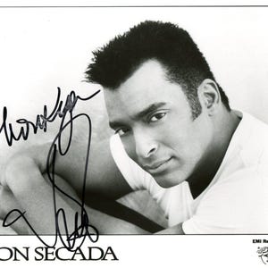 May include: Black and white photograph of Jon Secada, with his signature. He is wearing a white shirt and looking towards the camera. The text "JON SECADA" is at the bottom of the image.
